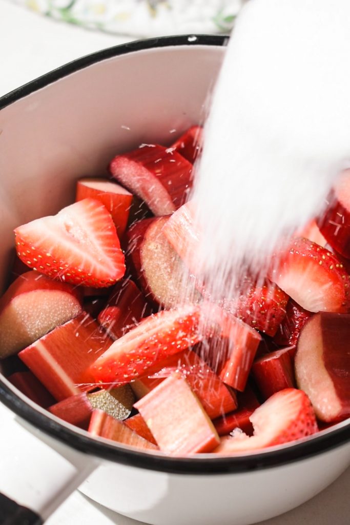 Adding sugar to fresh strawberries and rhubarb in a saucepan.