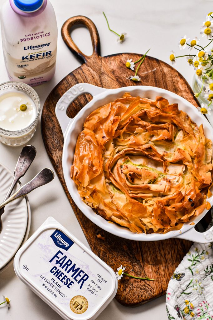Farmer cheese phyllo pie in a white pie dish next to a glass of kefir and a tub of farmer cheese.