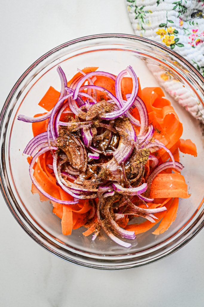 Carrot ribbons, sliced onions and spices in a glass mixing bowl.