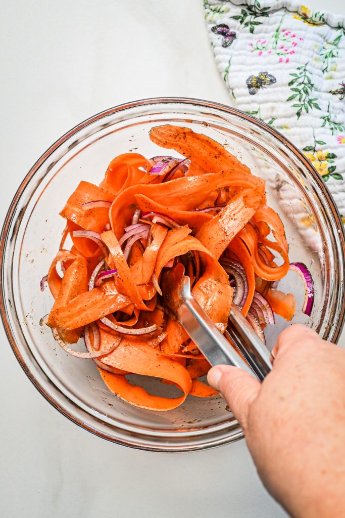 Using tongs to mix together the carrot ribbons, onions and spices.