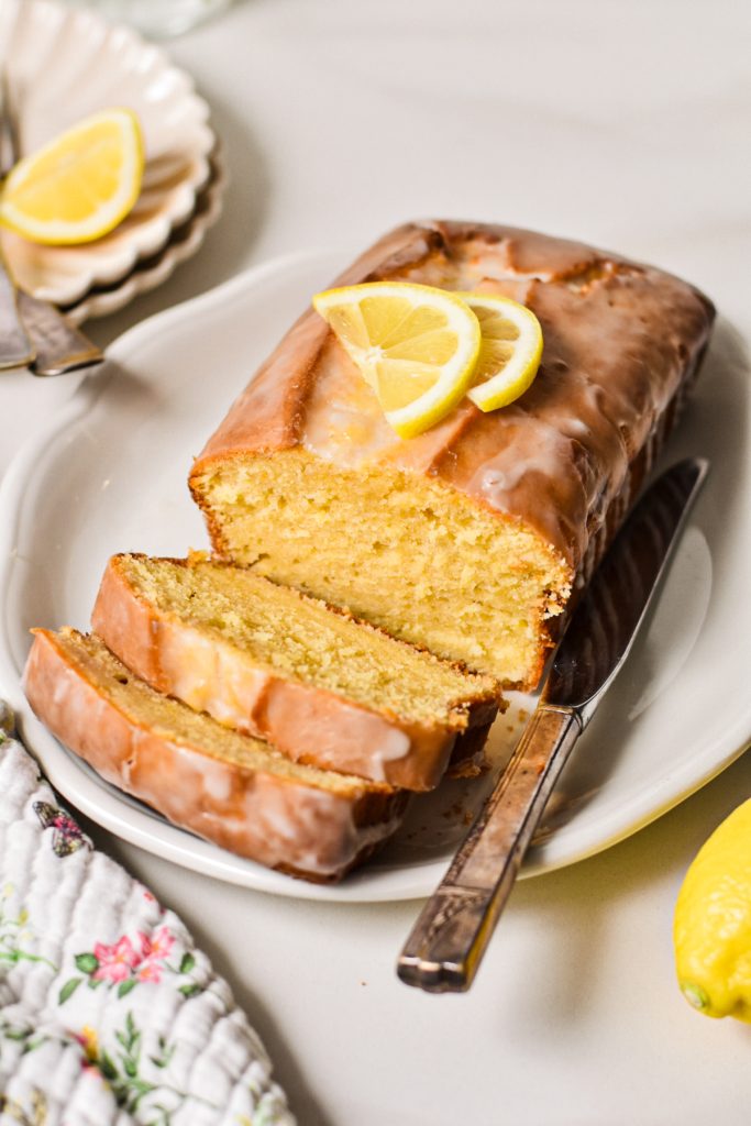 Lemon loaf cake with a glaze and fresh lemon on top with two slices off the end.