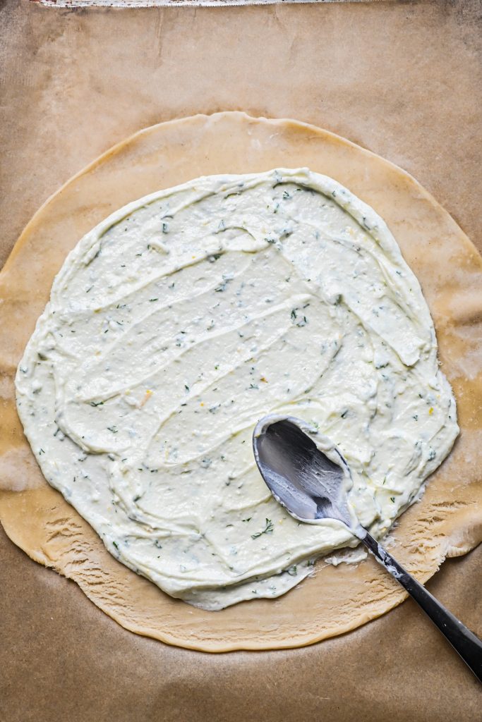 Using a spoon to spread the feta filling onto a pie crust on parchment paper.