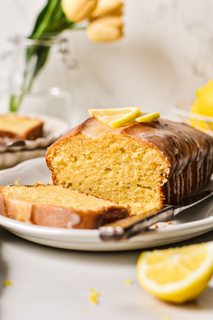 Lemon loaf cake on a plate with a slice off the end and fresh lemon on the top.