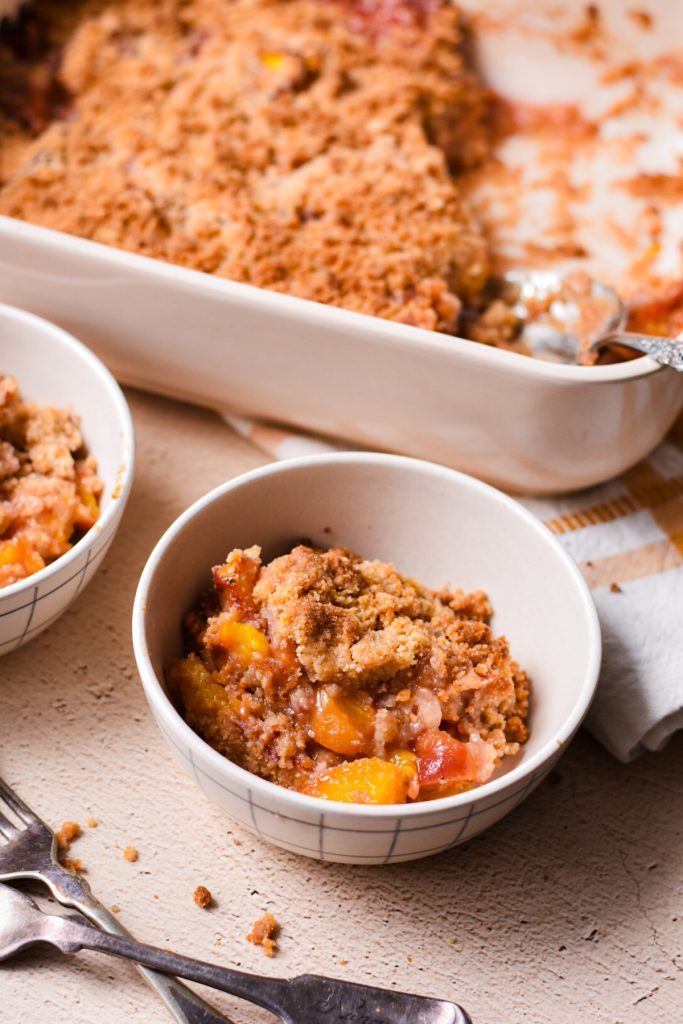Two bowls with peach crisp in them with the baking dish behind them.