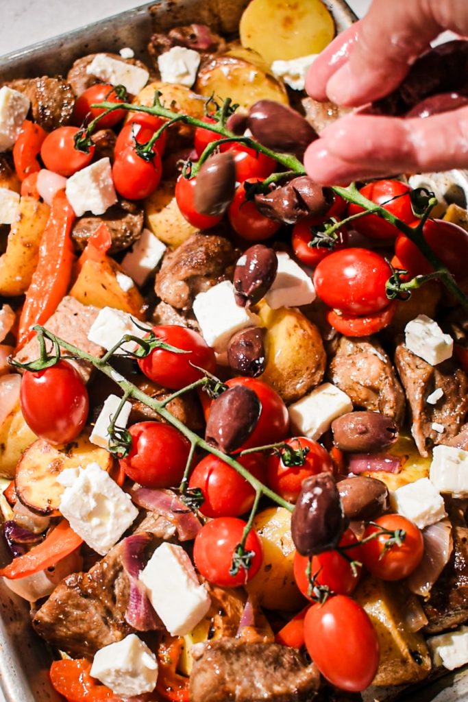 Hand placing the tomatoes, olives and feta on top of the roasted pork and veggies.