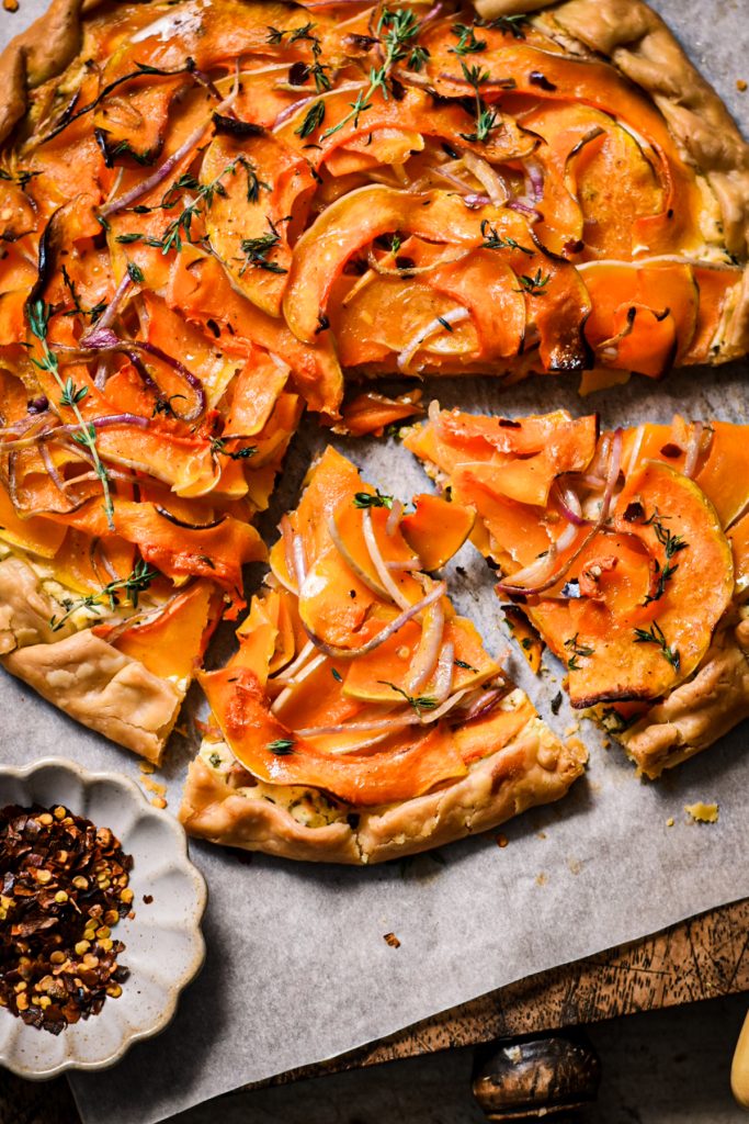 Butternut squash galette with two slices missing next to a small bowl of chili flakes.