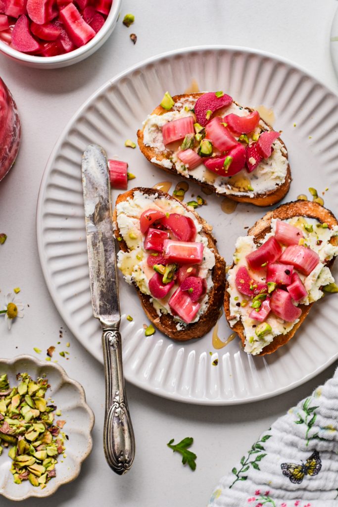 Toast with cream cheese, pistachios, pickled rhubarb and honey served on a plate.
