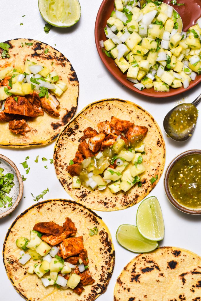 Chicken al pastor tacos assembled on a white background next to the pineapple pico de gallo.