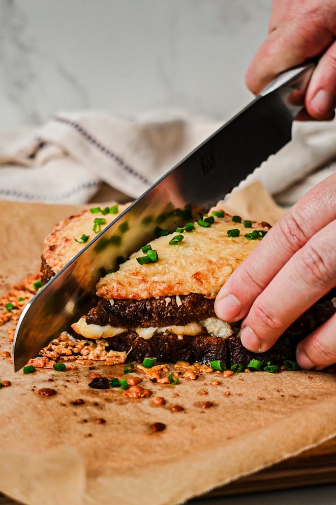 Hand slicing the croque monsieur sandwich on a sheet pan.