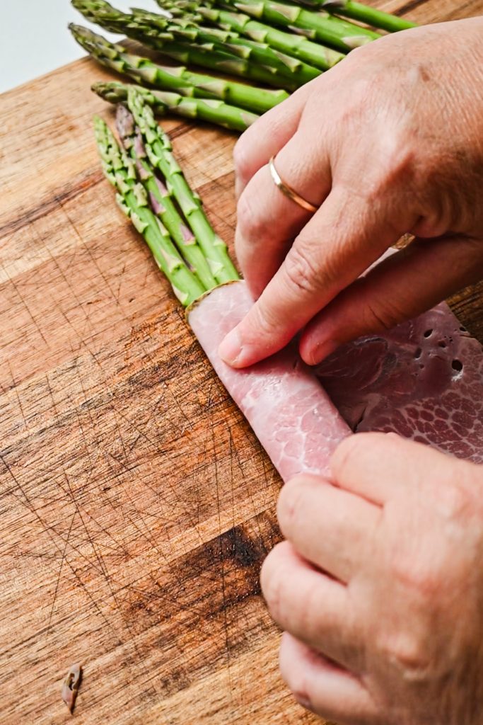 Hands wrapping asparagus with ham on a wooden board.