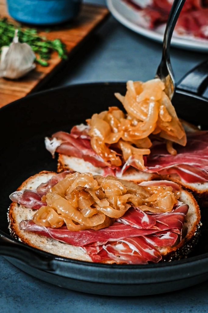 Adding caramelized onions on top of the the prosciutto layer on the bread.