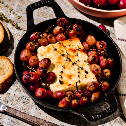 Block of baked feta in a cast iron skillet surrounded by baked grapes.