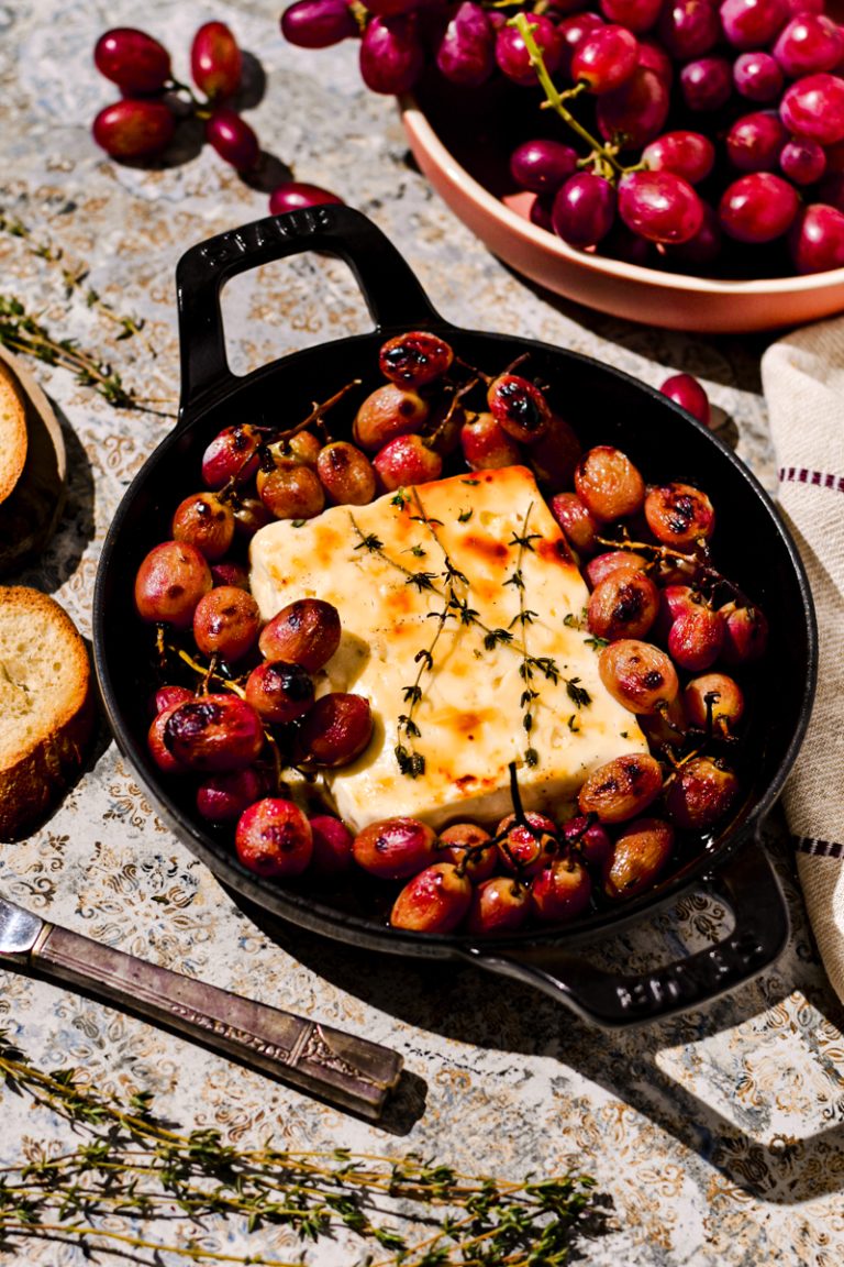 Block of baked feta in a cast iron skillet surrounded by baked grapes.