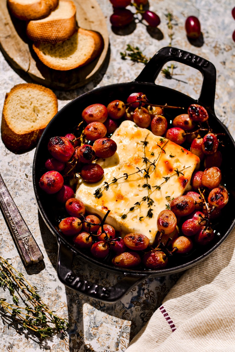 Baked feta with honey and grapes in a cast iron skillet next to some toasted bread.