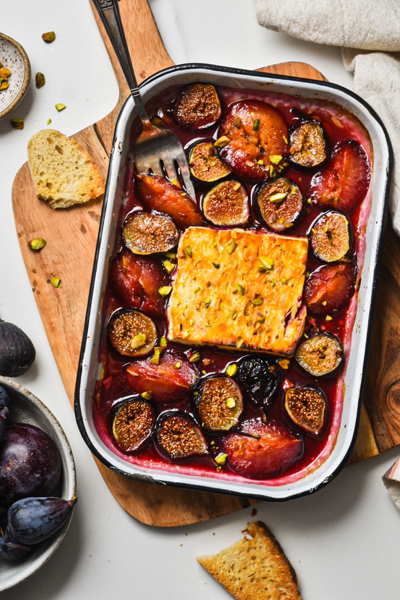 Overhead shot of baked feta with figs and plums in a baking dish on a wooden board.