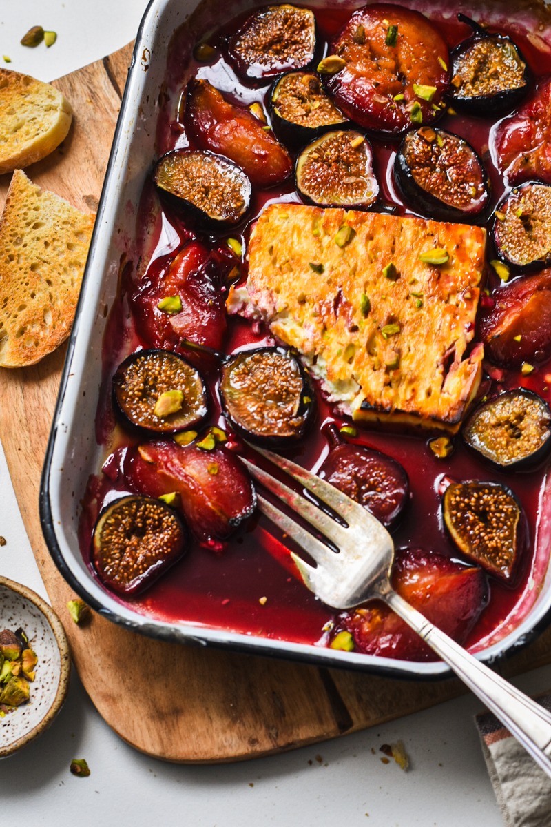Baked feta with figs and plums in a baking dish next to some toasted bread.