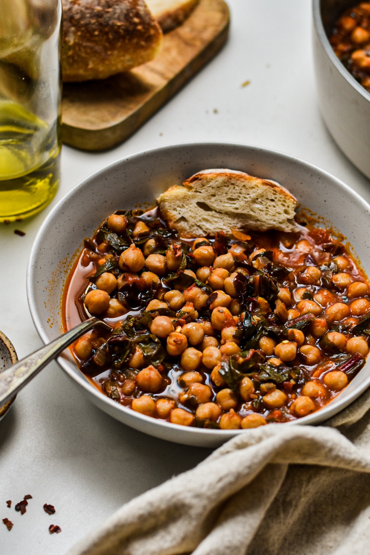Grey bowl with chickpea and Swiss chard soup served with a spoon and bread on the side.