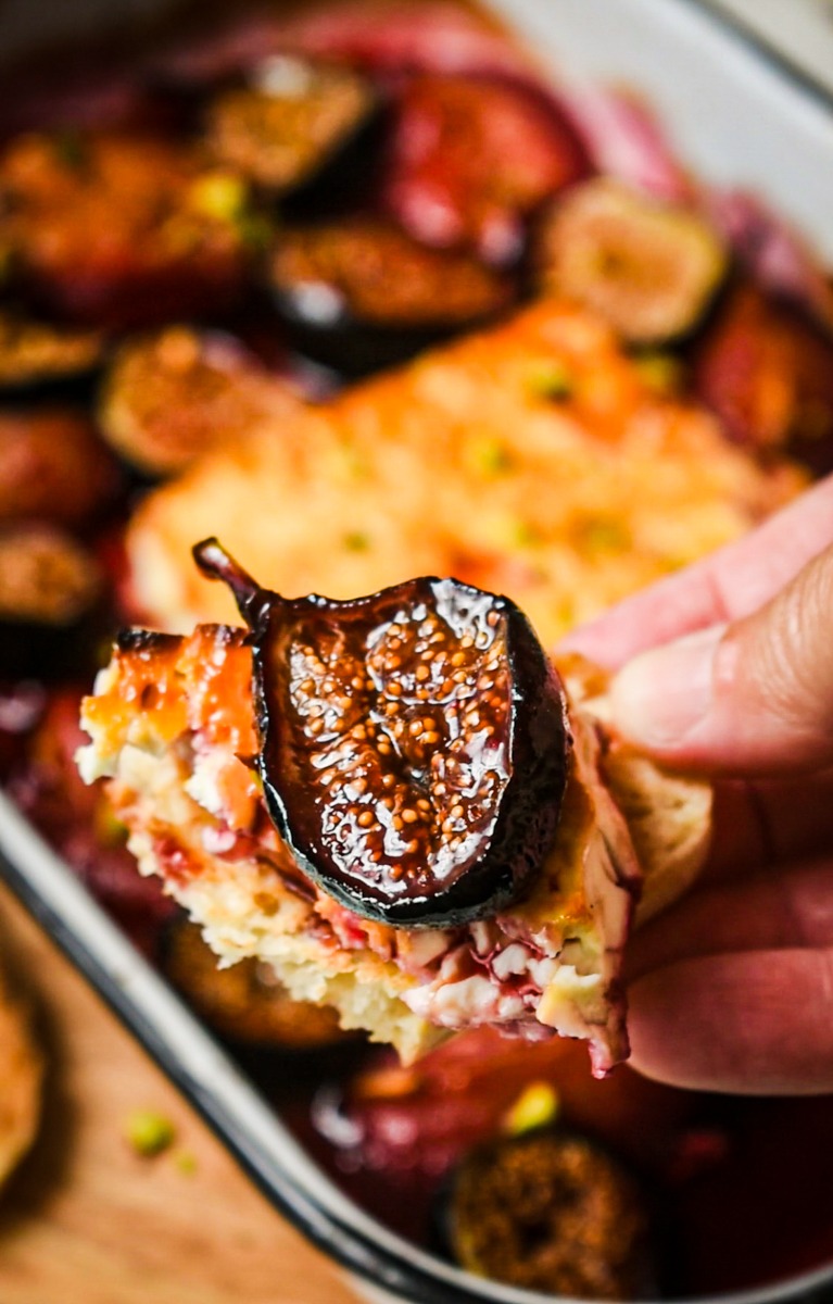 Hand holding a piece of bread with baked feta and a fig on top.