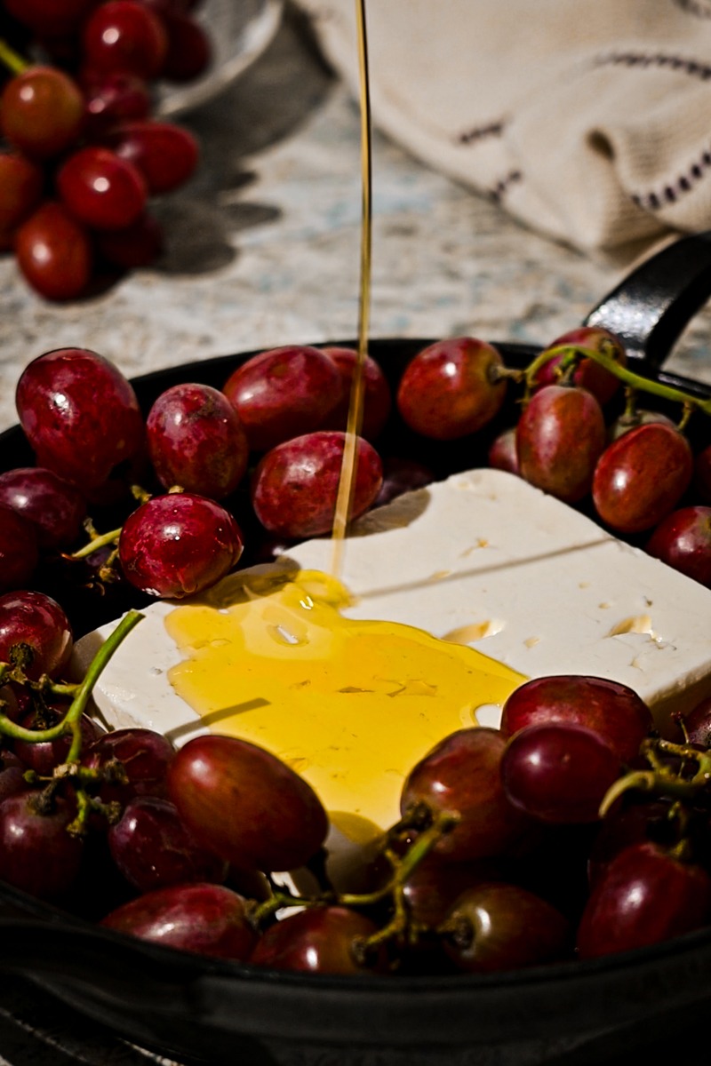 Block of feta in a cast iron with grapes with olive oil being poured on top.