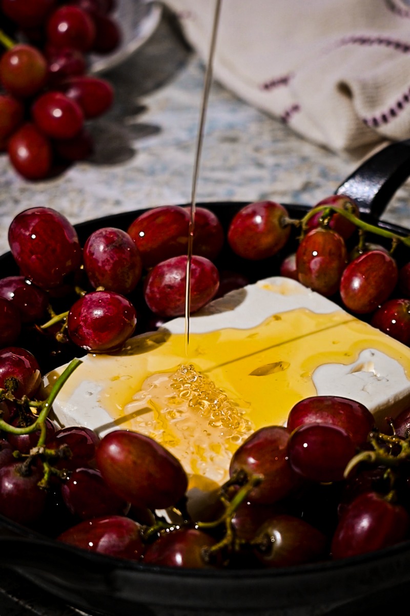 Honey being poured on top of the block of feta surrounded by grapes.