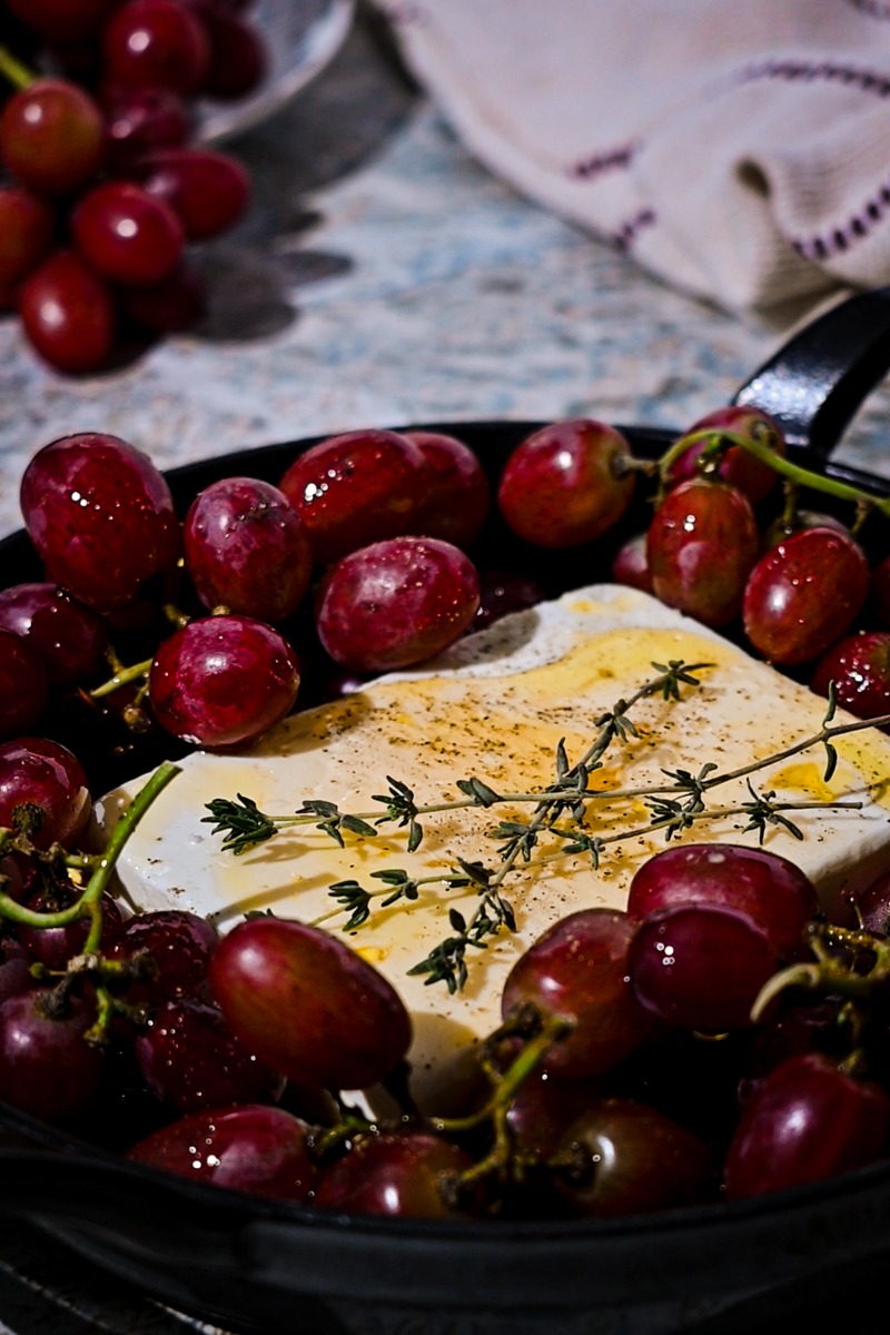 Fresh sprigs of thyme placed on top of the block of feta that is surrounded by grapes.