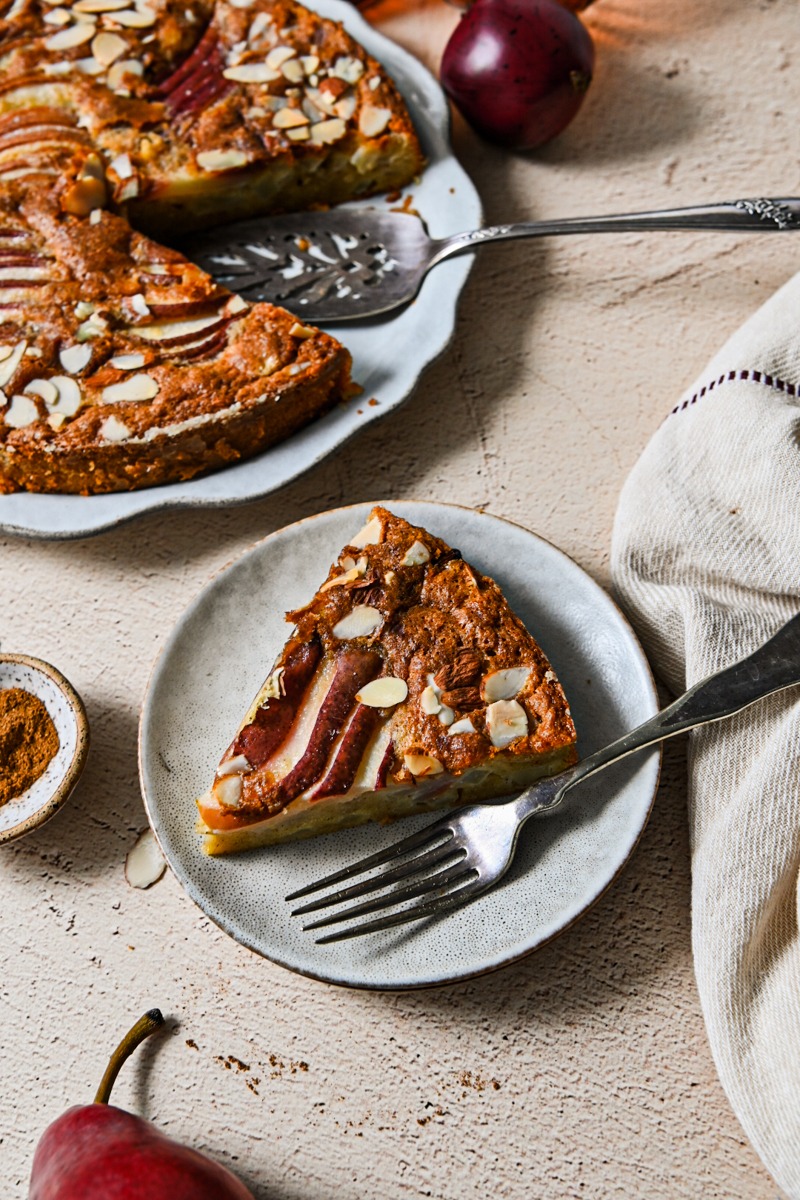 Slice of spicy pear French cake on a plate with the rest of the cake behind it.