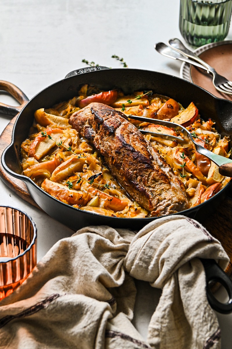 Side view of a cast iron skillet with cabbage and apples and a pork loin in the middle.