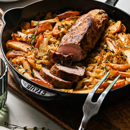 Pork loin with some slices off of the end on a bed of cabbage and apples in a skillet.