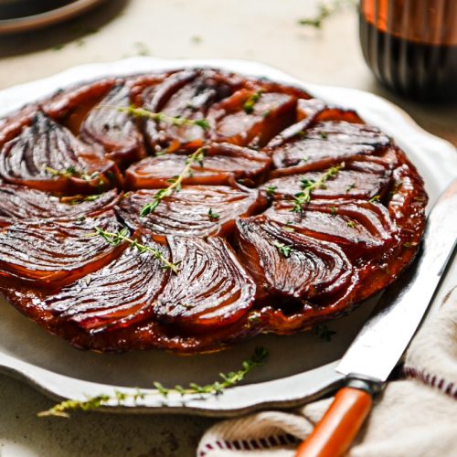 Red wine onion tart served on a platter with fresh thyme garnishing on top.
