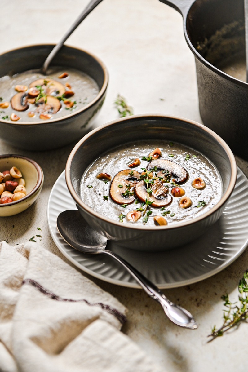 Two bowls of creamy mushroom soup with one of them served on a plate with a spoon.