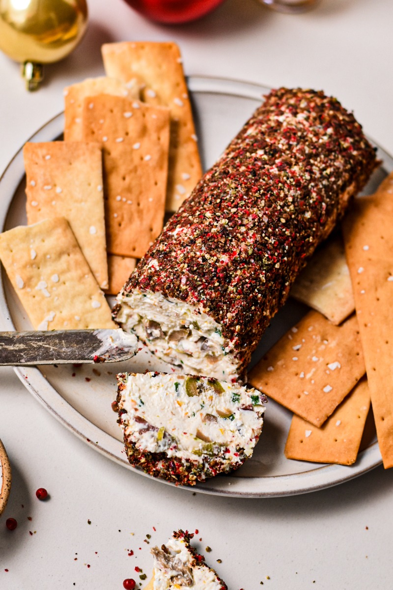 Olive and feta cheese log coated in pepper served on a plate with crackers.